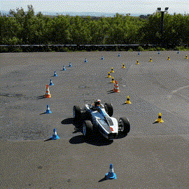 An autonomous electric vehicle with no driver navigates a cone-marked track while being followed by a drone.